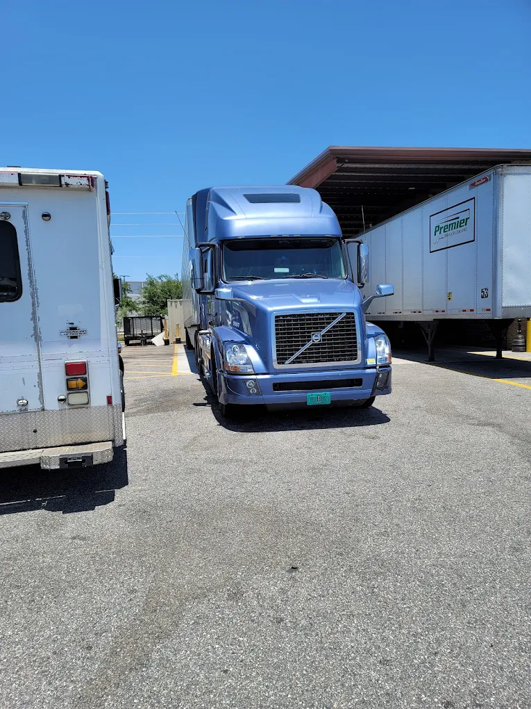 Blue Volvo semi truck parked between white trailers in a lot under clear sky